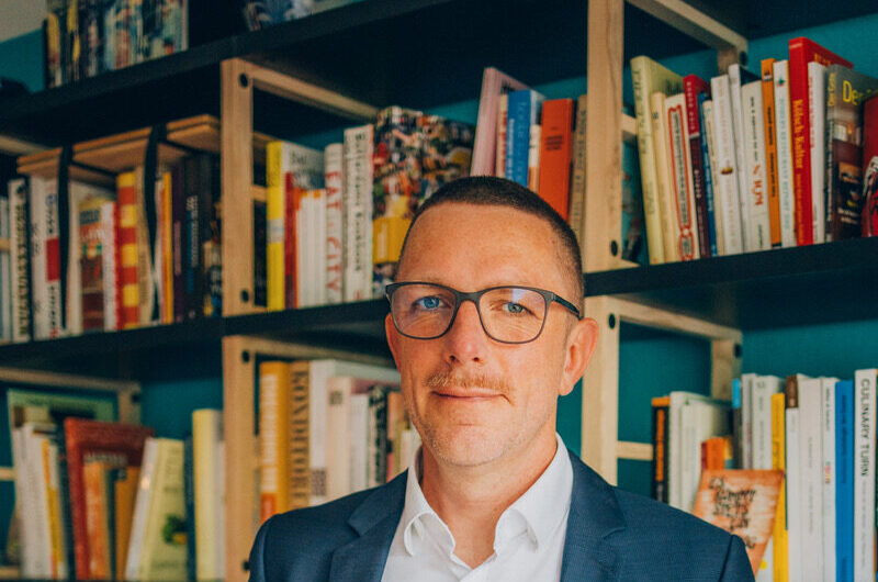 Porträt Johannes J.Arens im Anzug vor einem Bücherregal.