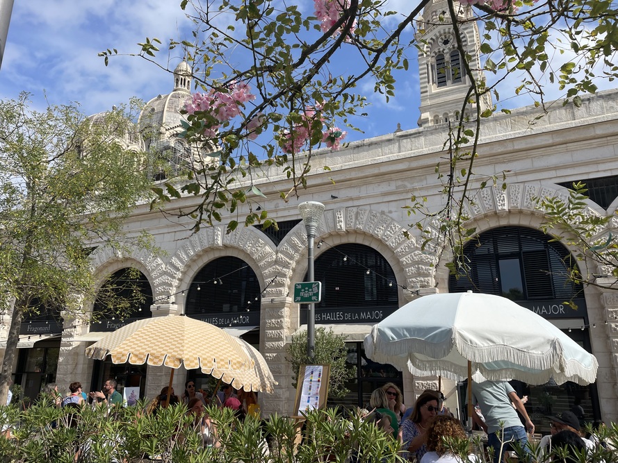 Straßencafé vor Les Halles de la Major in Marseille – Menschen unter Sonnenschirmen, Kulisse mit historischen Arkaden